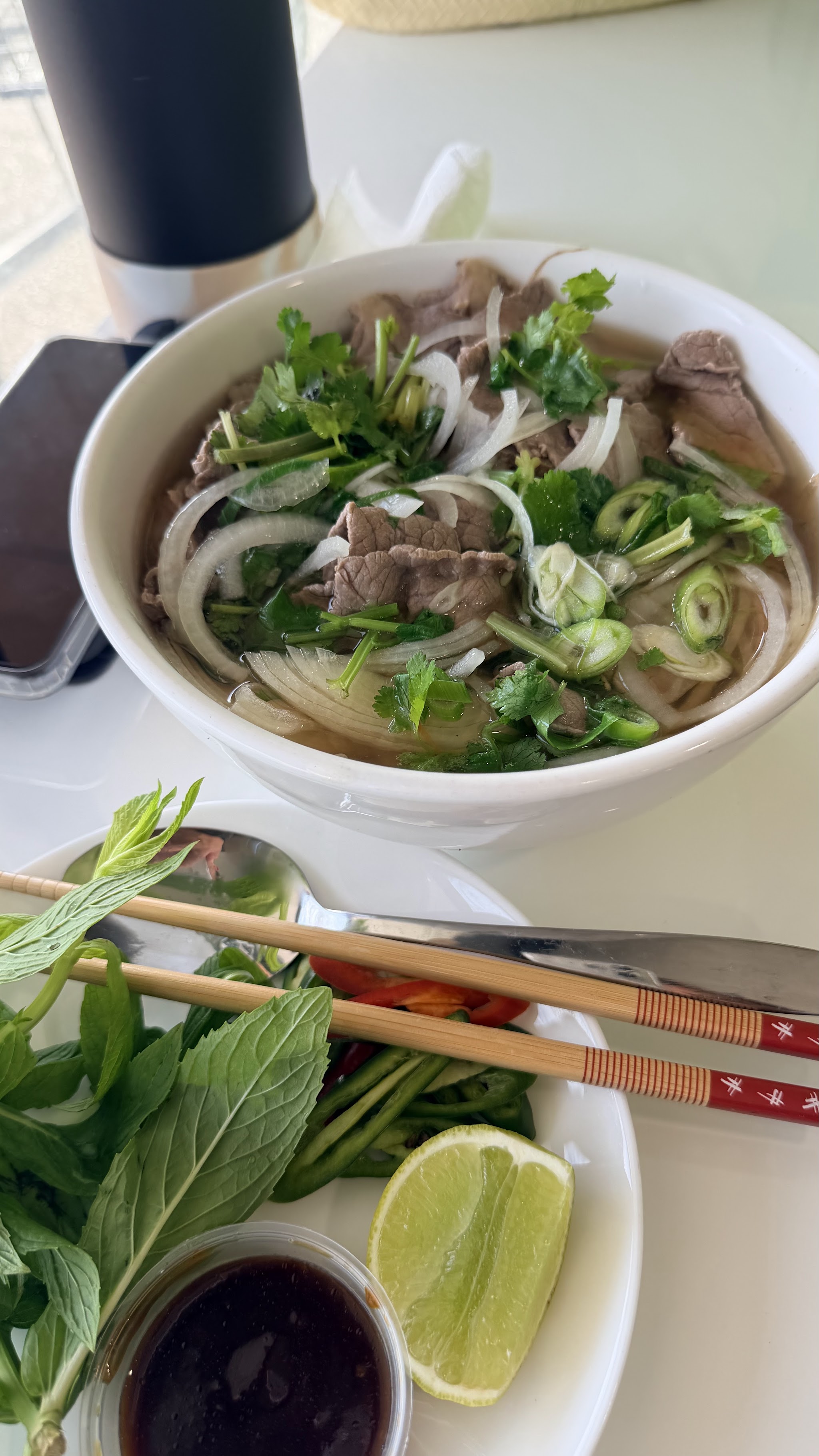 A bowl of beef phở with sliced beef, onion and herbs, beside a plate of fresh garnish